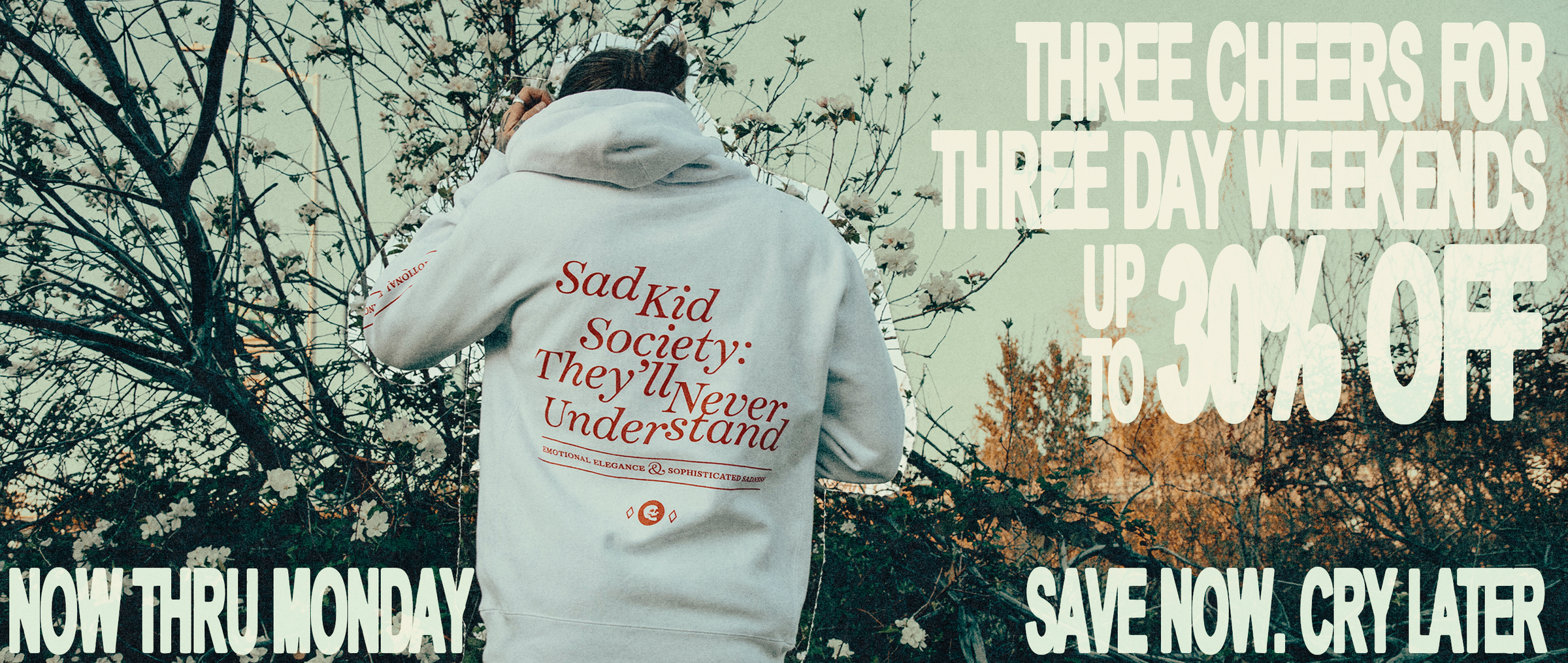 A person viewed from behind wearing a white hoodie with red text reading 'Sad Kid Society: They'll Never Understand' outdoors among flowering trees. Large promotional text overlays read 'THREE CHEERS FOR THREE DAY WEEKENDS UP TO 30% OFF', 'NOW THRU MONDAY', and 'SAVE NOW.